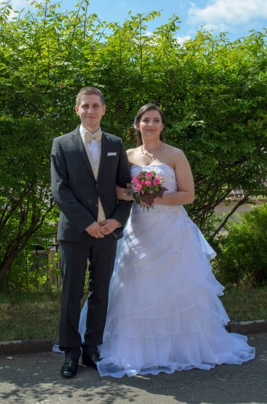 Hand in Hand ins Glück Braut im Hochzeitskleid mit Brautstrauß neben ihrem Bräutigam vor grüner Landschaft.