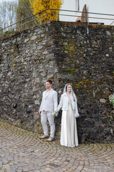 Zwischen zwei Wegen Ein Brautpaar steht an der Ecke einer Steinmauer, beide in eleganter Hochzeitskleidung und halten sich fest an den Händen.