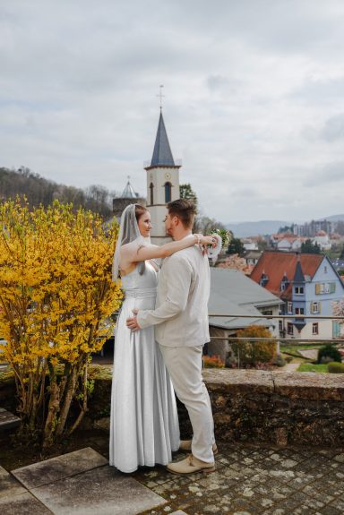 Blühende Liebe Brautpaar umarmt sich vor einer Kirche, umgeben von herbstlichen Bäumen und Häusern.