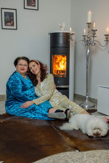 Zwei Frauen in eleganter Kleidung sitzen gemütlich vor einem Kamin, ein Hund liegt neben ihnen.
