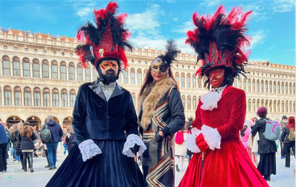 Drei Personen in bunten Kostümen und Masken vor historischer venezianischer Architektur.