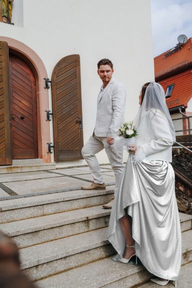 Der Weg zur Kirche Brautpaar auf einer Treppe, Braut mit Blumenstrauß, beide in eleganter Hochzeitskleidung.