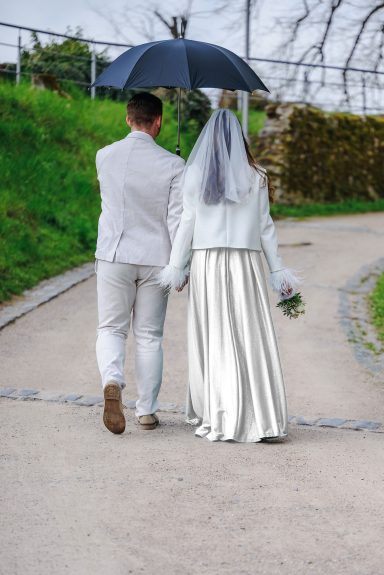 Liebe kennt kein Wetter Ein Brautpaar geht Hand in Hand einen Weg entlang, unter einem Regenschirm.