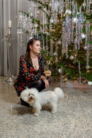Frau mit eleganter Kleidung sitzt neben einem kleinen Hund vor einem festlich geschmückten Baum.